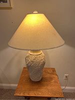 Cream-colored ceramic table lamp with textured vase-shaped base and conical lampshade on a wooden surface against a neutral wall.