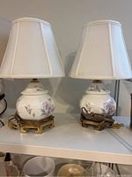 Pair of small ceramic floral table lamps with white shades shown on a shelf.
