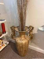 Large gold-tone metal urn with ornate handles and vertical ribbing, filled with tall dried reed stems.