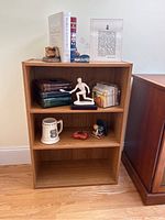Photo of first wooden bookcase with three shelves, displaying various books and decorative items, contents not included.