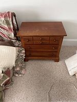 Front view of one Stanley night table showing three small upper drawers and one larger bottom drawer with brown wood finish, placed on carpet near chair and blanket.