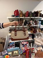 Photo showing a beige and brown structured handbag being held in front of shelves full of various handbags and purses.