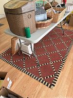Rug laid out flat on hardwood floor with various baskets and boxes on a folding table above it, showing the rug's size and pattern.