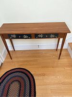 Photo showing a wooden side table with two drawers decorated with floral painted designs on fronts, on a wooden floor near a white wall and a patterned rug.