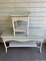 Two white wicker tables stacked: small square end table on top of larger rectangular side table, showing wicker texture, construction, and dimensions relative to porch background