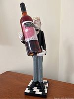 Side front view of wine butler figurine holding an empty wine bottle placeholder, on a checkered base.