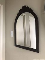 Wall-mounted antique mirror showing dark wooden frame with carved scroll detail at top, rectangular arched shape, reflecting door and light switch.