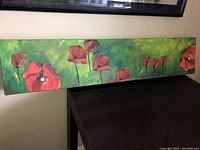 Front view of the long rectangular canvas art of red poppies on green background resting on a dark wood surface.