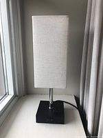 Full view of bedside lamp showing white rectangular fabric shade, chrome stem, and black base against wall and window.