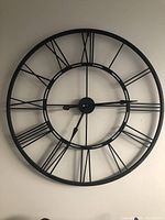 Front view of large black metal wall clock showing Roman numerals and black hands on a wall.