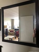 Front view of the square beveled mirror in a dark wood frame reflecting a living room with furniture.