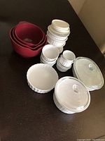 Overall view of the white ceramic bowls and casseroles with glass lids, plus red mixing bowls on table.