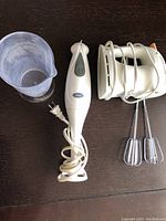 Oster immersion blender, power cord coiled, two metal beaters with white attachments, plastic mixer stand, and clear plastic mixing cup.
