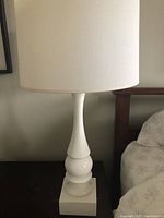 Front view of the modern white ceramic bedside lamp with off-white fabric shade standing on dark brown bedside table next to bed.