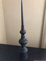 Tall black decorative obelisk photographed against a plain wall and placed on a black surface, showing its full height and detailing.