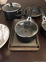 Cookware set displayed including various pots, pans, lids, steamer basket and baking sheets on dark tabletop