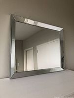 Photo showing a rectangular silver framed mirror with beveled edges, placed against a wall reflecting a room corner.