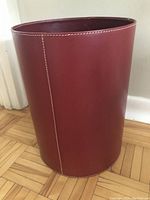Side view of red cylindrical waste paper basket showing leather-like texture and stitching detail.