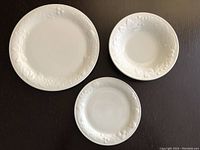 White dinner plates, side plates, and bowls showing embossed floral rims on white ceramic.