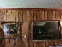 Both framed paintings mounted on wood-paneled wall, left smaller interior scene and right larger forest landscape