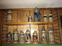 Wooden shelf showing five ceramic steins on upper shelf and six on lower shelf including clear glass stein.