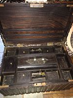 Photo showing open carved wooden chest with hinged lid lifted, displaying multiple small drawers and compartments inside, some with metal knobs, and a removable center section with wear marks.