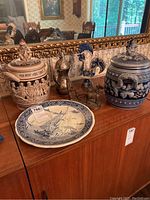 Photo showing two ceramic covered urns with detailed bas-relief figurative scenes, one with a seated gnome figure on the lid, along with a Delft-style ceramic platter, ceramic rooster figurine, metal pitcher and metal oxcart figurine on wooden cabinet.