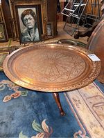 Copper table showing etched round tabletop with intricate patterns on copper surface, sitting on dark wood folding base with claw feet. Background includes paintings and other decorative items.
