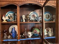 Full view of wooden cabinet displaying multiple figurines, art glass vases, decorative plates, a rooster figurine, stemware, and a pitcher, showing the arrangement of the lot contents.