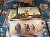 View of several framed paintings laid on the floor, showing at least four different artworks including a religious figure painting and landscape themes