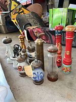 Wide view showing a set of 10 assorted pepper mills of mixed materials ceramic, glass, metal, and wood arranged on a concrete floor near boxes.