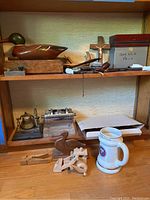 Shelf with a brass bell on wooden base, wooden duck decoy, various smoking pipes, spark plug cutaway model, wooden boxes, courtroom gavel, and ceramic mug.