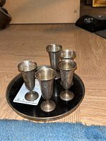 Five tarnished sterling silver cordial cups arranged on a small round tray on a wood floor.