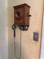 Front view of entire wooden wall telephone mounted on wall showing mouthpiece, hand crank and bells