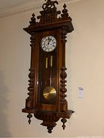 Full view of carved wood wall clock mounted on wall showing overall design, carved finials and crest.