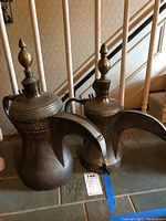 Photo of two metal pitchers showing general size, shape, curved handles, long spouts, and finial-topped lids with decorative etchings