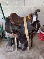 Photo showing full side view of the western saddle with wooden stirrups and beige girth strap on a stand.