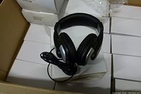 Single Synchrotech GC-MM-HEADPHONE over ear stereo headphone placed on a white box inside a cardboard box. Headphones have black cushioned ear pads, silver accents, and attached audio cable.