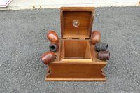 View of the wooden tobacco pipe stand with hinged lid open showing internal compartment and six assorted vintage pipes around the stand.