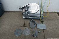 Accessories including multiple perforated metal plates, meat grip, and additional attachments spread on the floor in front of the slicer.