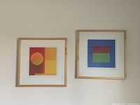 Two square wooden framed abstract art pictures hanging on a light wall surface, showing color blocks in warm sunset tones on the left, and cooler geometric color blocks on the right.
