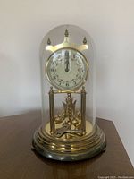Brass anniversary mantel clock under a glass dome with white dial and Arabic numerals, on round brass base and winding key in front.