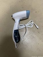 Handheld clothes steamer lying on flat surface showing front, side, water reservoir, and attached power cord.