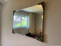 Photo shows a rectangular wall mirror mounted on a light-colored wall. Mirror has beveled edges and decorative gold corner ornaments with intricate scroll and floral design. Reflects window, curtain, chair, lamp and floral arrangement.