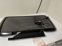 Black rectangular electric hot plate cooking griddle with power cord and temperature control knob on a white table with power cable coiled on top.