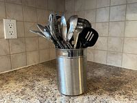 Photo of a stainless steel container holding assorted metal kitchen utensils on a kitchen counter