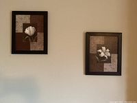 Two matching framed wall pictures featuring white flowers on geometric colored backgrounds hung on a wall.