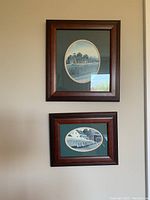 Two framed farm pictures by Catherine Karnes Munne Collection hanging on a wall. The top picture is larger with an oval farm scene, the bottom one smaller also with an oval farm scene. Both frames are wood with a dark green matte.
