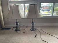 Photo of two matching table lamps with brass look bases and beige bell-shaped fabric shades, placed on carpet near window with cords visible.