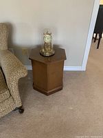 Angle front view of the octagon-shaped wooden side table with a retro style, placed next to a beige textured armchair, showing good condition and natural wood grain.
